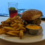 Sezamový burger s hovädzím mäsom a hranolkami na bielej tanieri, podávaný s miskou oranžovej omáčky, v pozadí rozmazaný pohár s červeným nápojom a mätou.