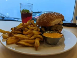 Sezamový burger s hovädzím mäsom a hranolkami na bielej tanieri, podávaný s miskou oranžovej omáčky, v pozadí rozmazaný pohár s červeným nápojom a mätou.