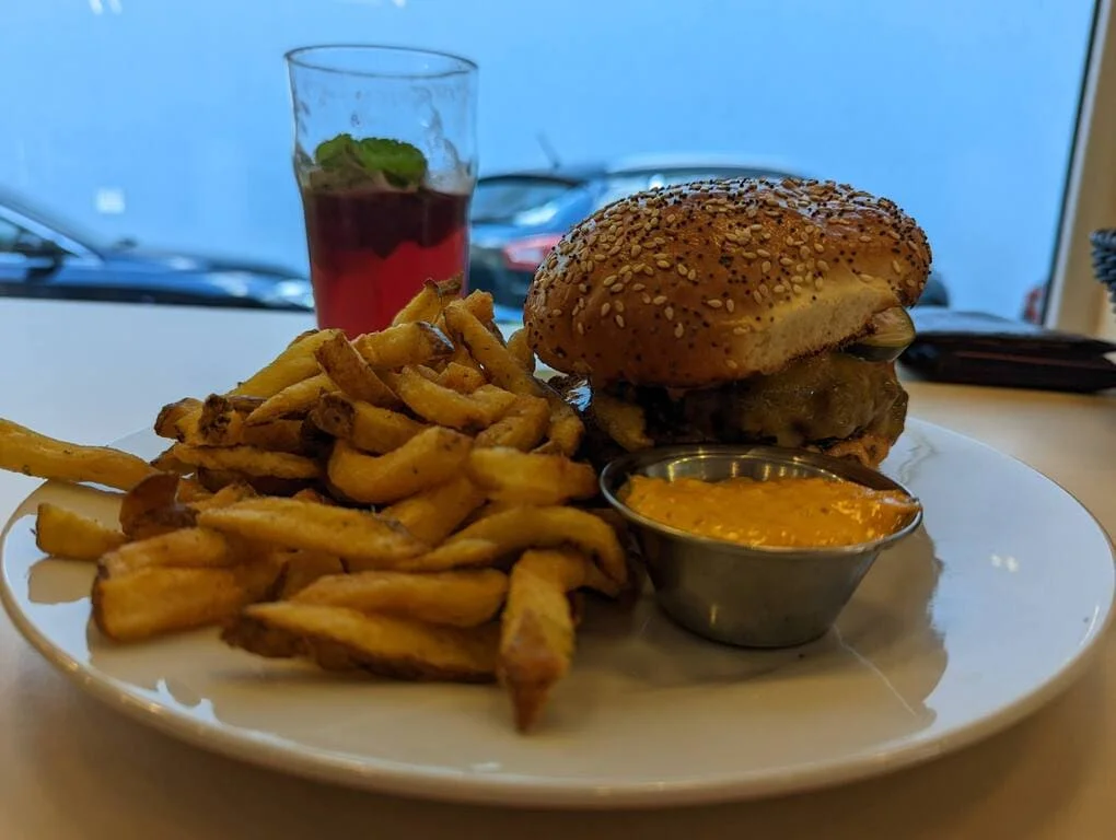 Sezamový burger s hovädzím mäsom a hranolkami na bielej tanieri, podávaný s miskou oranžovej omáčky, v pozadí rozmazaný pohár s červeným nápojom a mätou.