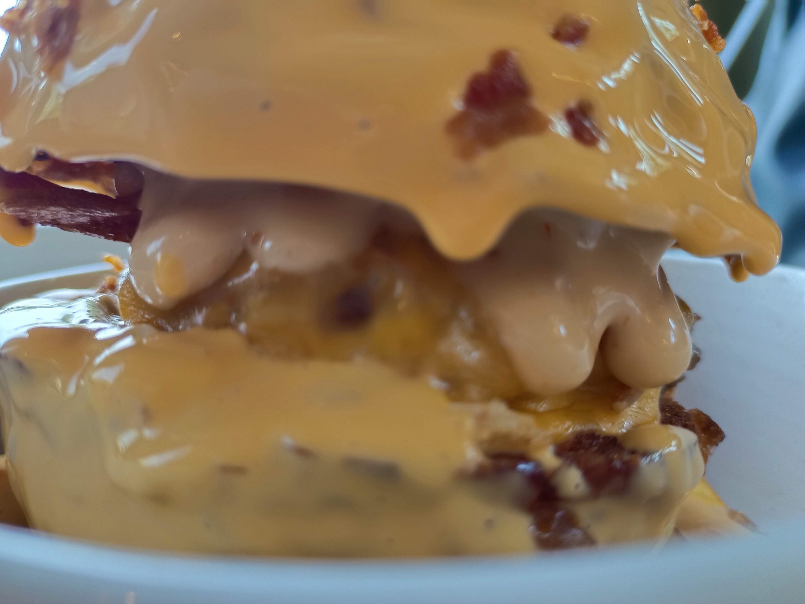 Detailný záber na burger s vrstvami roztopeného syra a slaniny, zaliaty bohatou syrovou omáčkou, servírovaný v bielej porcelánovej mise.