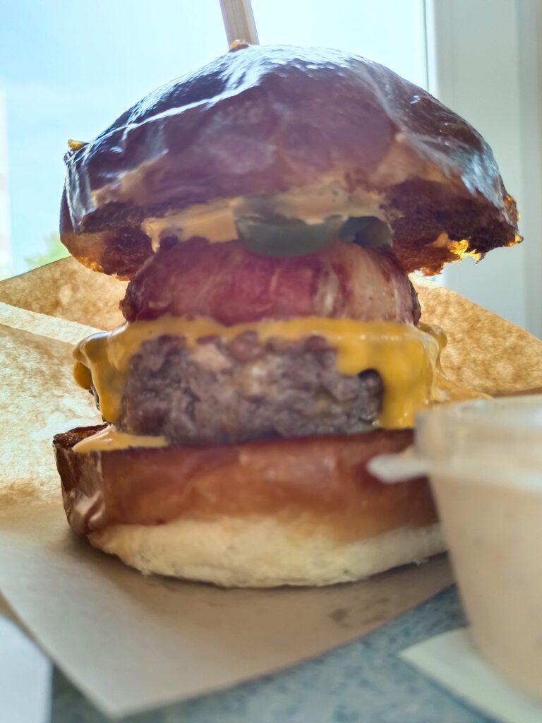 Zblízka pohľad na burger s tmavou žemľou, hovädzím mäsom, syrom a kyslou uhorkou, podávaný s malým pohárom omáčky na pergamene nad bielou doskou.