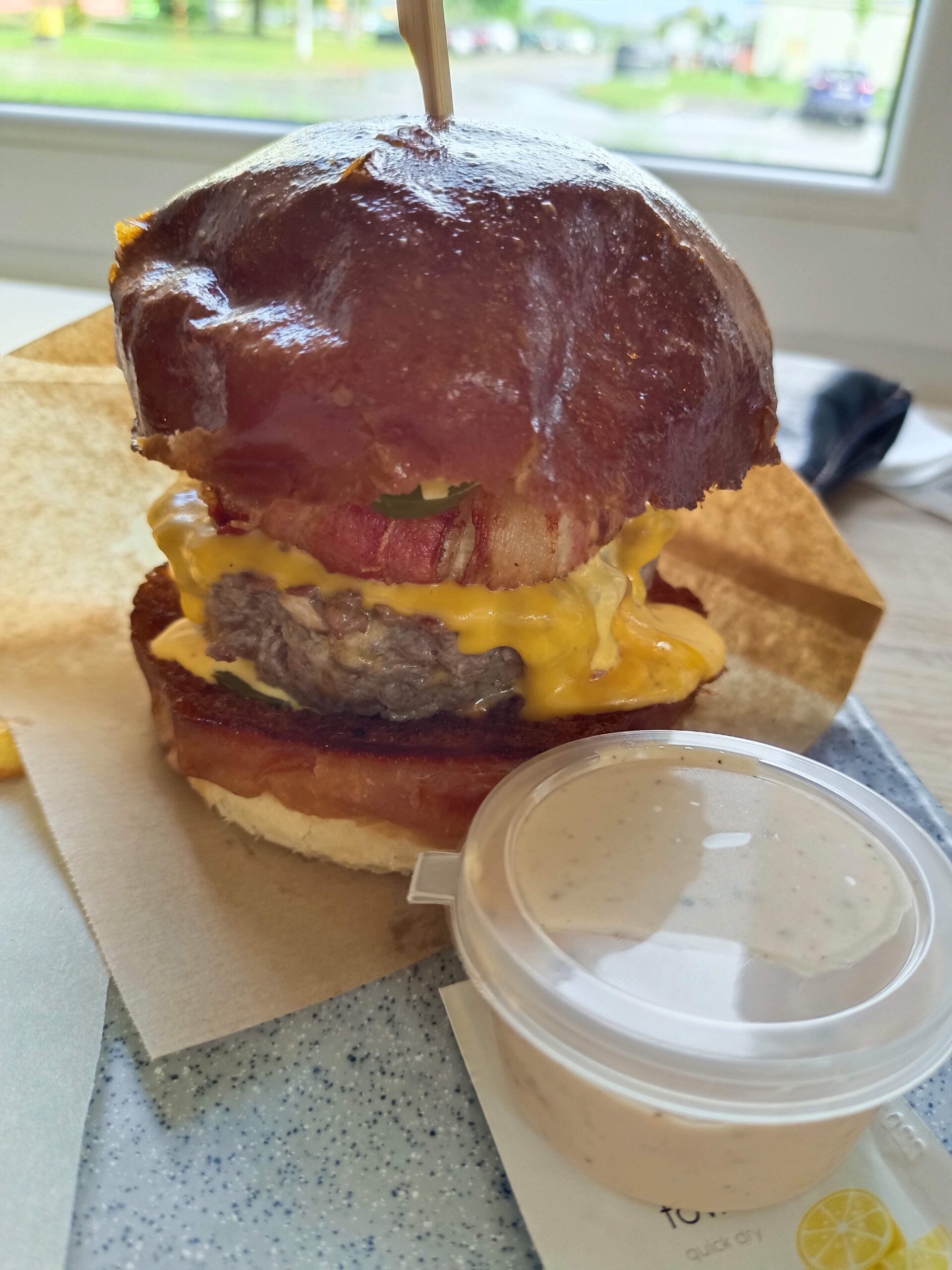 Burger s lesklou žemľou, syrom, slaninou a uhorkou, zastrčený dreveným špajdlou, servírovaný s pohárom bielej omáčky na svetlom stole pri okne.