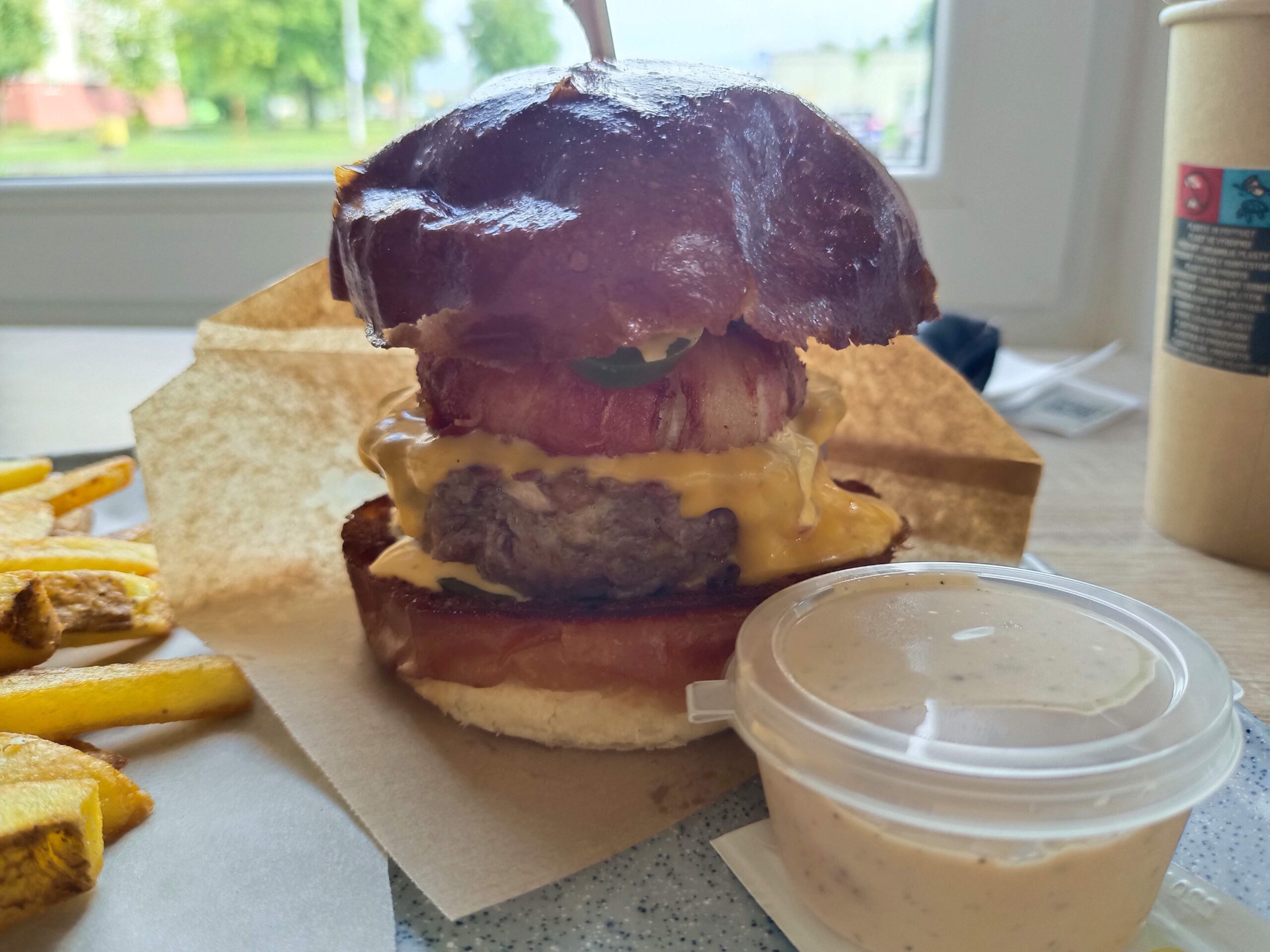 Burger s tmavou žemľou, vrstvou rozpusteného syra, slaninou a zeleninou, podávaný s hranolkami a miskou ranch omáčky na svetlom stole pri okne.