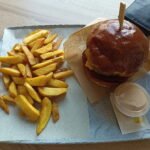 Burger s hnedou žemľou, syrom a plátkom mäsa podávaný na papierovej podložke spolu s porciou hranolčekov a miskou bielej omáčky na keramickej tanieri.