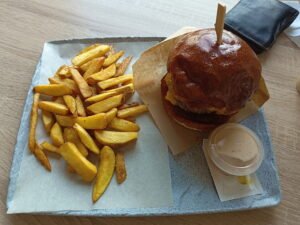 Burger s hnedou žemľou, syrom a plátkom mäsa podávaný na papierovej podložke spolu s porciou hranolčekov a miskou bielej omáčky na keramickej tanieri.