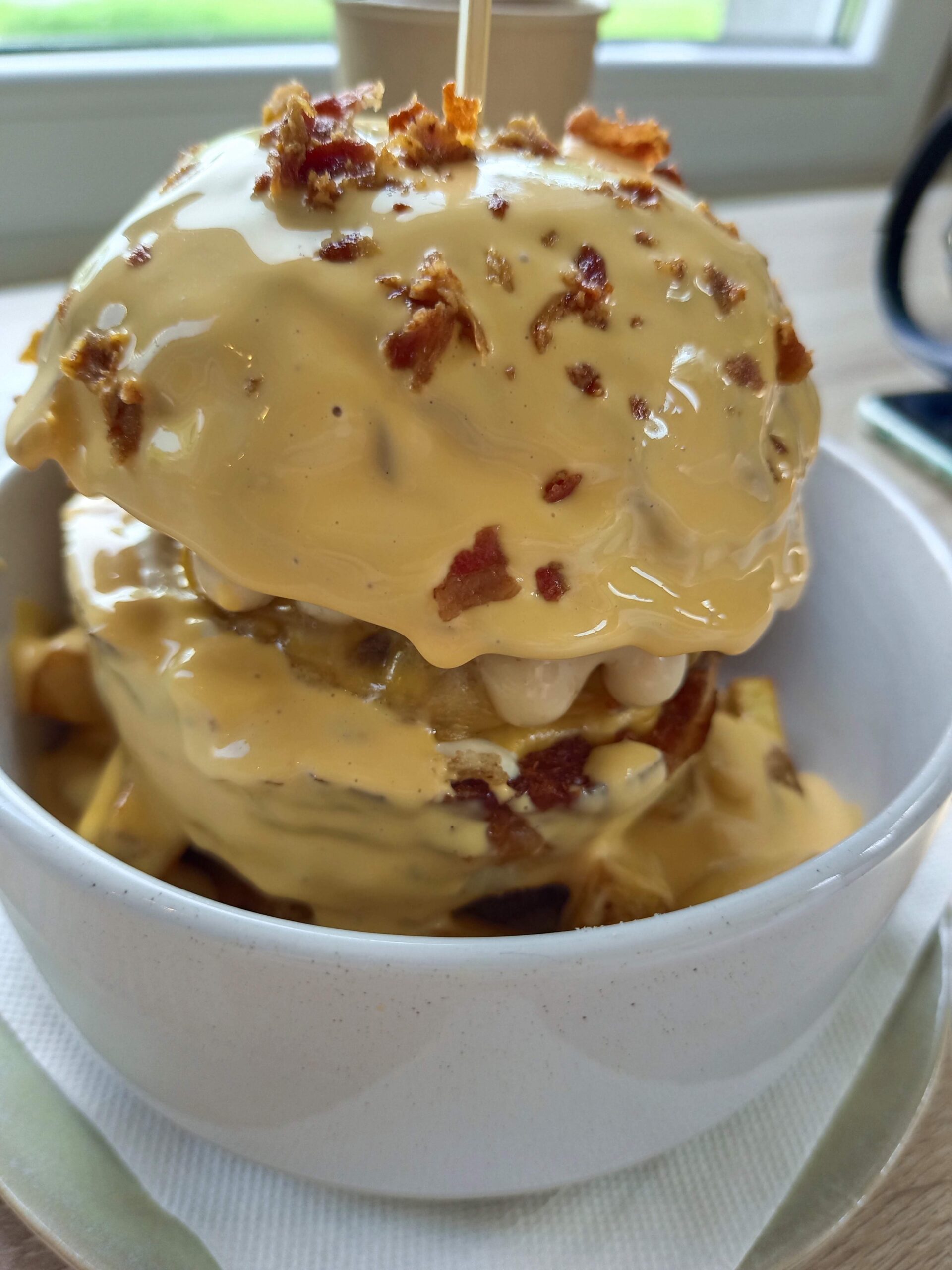 Burger s viacerými vrstvami, bohato pokrytý rozpusteným syrom a posypaný chrumkavou slaninou, podávaný v bielej porcelánovej mise.