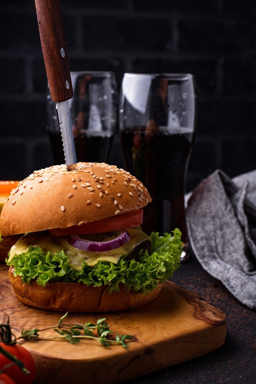 Burger so sezamovou žemľou, čerstvým šalátom, paradajkou a cibuľou na drevenej doske, do burgeru je zapichnutý nôž, v pozadí pohár tmavého nápoja a sivá utierka na tmavom pozadí.