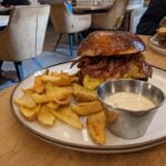 Sezamový burger so slaninou, syrom a hranolkami na bielej tanieri s miskou tatárskej omáčky, reštaurácia s retro dizajnom v pozadí.