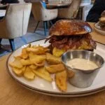 Sezamový burger so slaninou, syrom a hranolkami na bielej tanieri s miskou tatárskej omáčky, reštaurácia s retro dizajnom v pozadí.