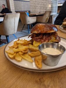 Sezamový burger so slaninou, syrom a hranolkami na bielej tanieri s miskou tatárskej omáčky, reštaurácia s retro dizajnom v pozadí.