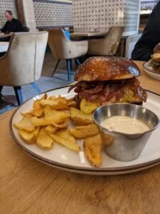 Sezamový burger so slaninou, syrom a hranolkami na bielej tanieri s miskou tatárskej omáčky, reštaurácia s retro dizajnom v pozadí.